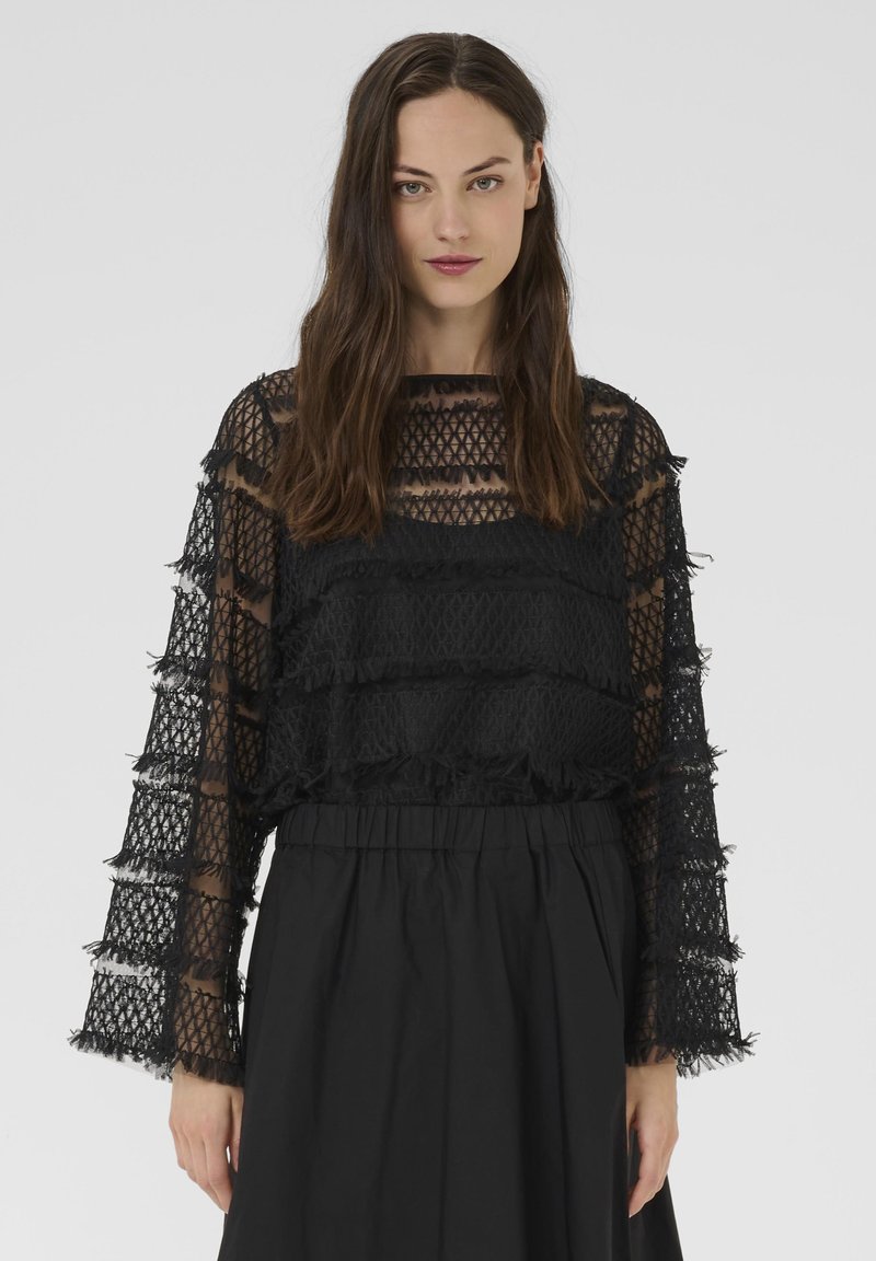 Sieviete ar gariem brūniem matiem, vilkusi melnu caurspīdīgu mežģīņu topu ar frizūru detaļām un augsta vidukļa melnām svārkiem uz vienkārša fona.