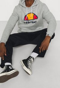 Sudadera con capucha gris con un logo gráfico en naranja, rojo y azul marino, combinada con pantalones de pana negros y zapatillas altas negras con detalles en blanco.