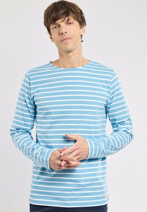 Homme aux cheveux châtain clair portant une chemise à manches longues à rayures bleues et blanches, debout les mains légèrement jointes.