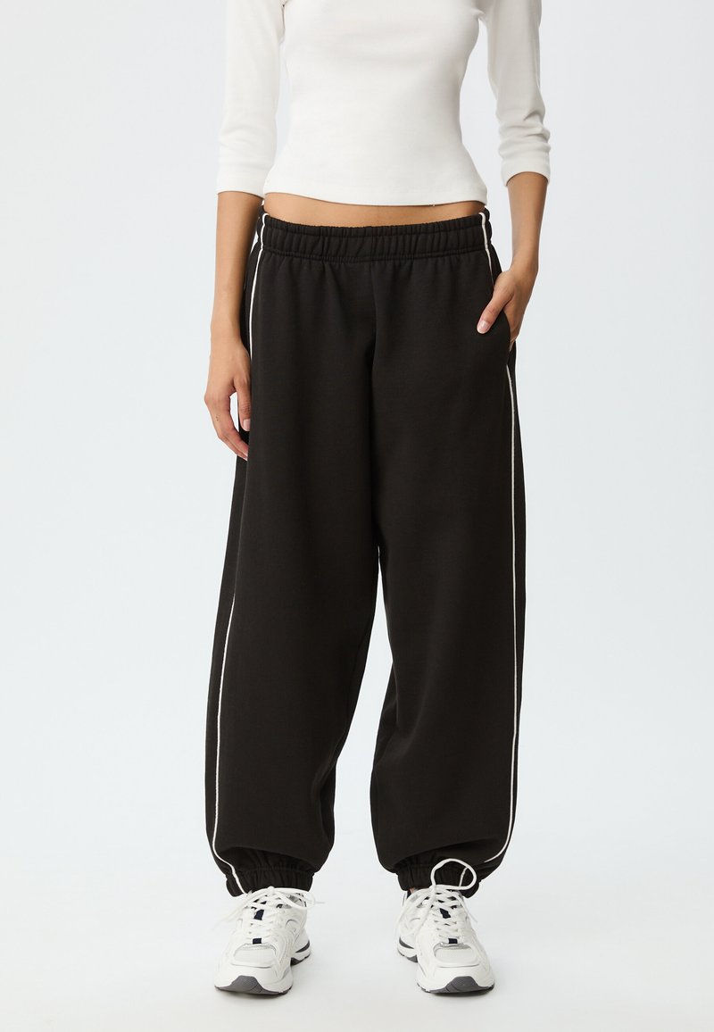 Zwarte trainingsbroek met elastische tailleband, witte zijstrepen en geribbelde enkels. Gemaakt van zacht stof, met een ontspannen, oversized pasvorm.