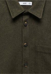 Olivengrøn uldskjorte med spredebånd, knaplukning foran og en enkelt brystlomme. Har sorte plastikknapper og en blød tekstur.