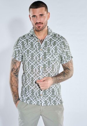 Mann mit kurzen Haaren und Tätowierungen auf beiden Armen, der ein grün-weiß gemustertes kurzes Hemd mit Knöpfen und helle Hosen trägt.