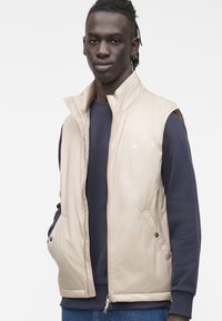 Bež prsluk bez rukava s visokim ovratnikom, izrađen od glatkog materijala. Ima prednji patentni zatvarač i bočne džepove sa zakovicama.