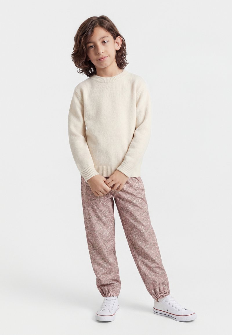 Dziecko o włosach do ramion, ubrane w kremowy sweter, różowe spodnie w wzory i białe trampki, stojące na jednolitym białym tle.