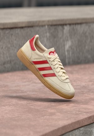 Beige suède sneaker met rode accenten, met drie strepen, een hartlogo op de hiel en een zool van natuurlijke rubber.