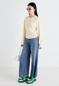 Top a maniche lunghe color crema, jeans di denim blu larghi, scarpe da ginnastica verde con plateau e borsa bianca. Il modello indossa occhiali da sole oversize chiari.
