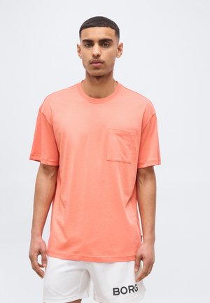 Άνδρας που φοράει κοραλί κοντομάνικο T-shirt με στρογγυλή λαιμόκοψη και τσέπη, καθώς και λευκά σορτς με το κείμενο "BORG" στο αριστερό πόδι, μπροστά σε λευκό φόντο.