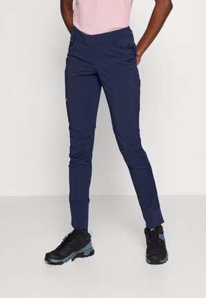 Persoon draagt marineblauwe slim-fit joggerbroek met gestructureerde manchetten en zwarte wandelschoenen, staat met handen in de zakken.