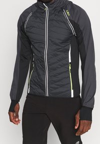 Veste noire et grise pour homme avec un design matelassé, dotée de fermetures éclair blanches et d'accents verts. Fabriquée en matériau léger et respirant.