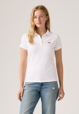 Femme aux longs cheveux blonds portant un polo blanc Levi's et un jean bleu clair, debout devant un fond uni.