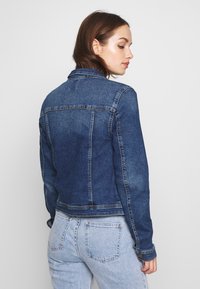 Denim jacket in dark blue, classic design with buttoned cuffs and two back seams. Styled with light blue jeans.