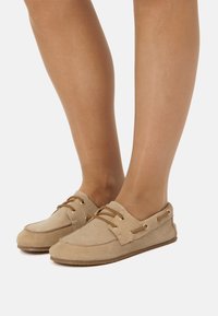 Zapatos náuticos de ante beige con cordones marrones, punta redonda y ojales laterales. Textura suave y suela plana, adecuados para un uso casual.