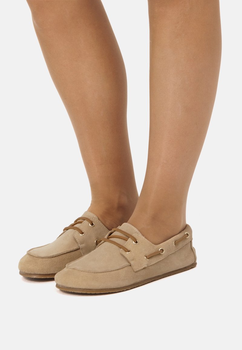 Zapatos náuticos de ante beige con cordones marrones, punta redonda y ojales laterales. Textura suave y suela plana, adecuados para un uso casual.