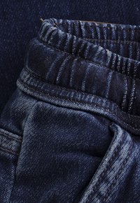 Zbliżenie na ciemnoniebieską tkaninę dżinsową, ukazujące elastyczny pas i szczegółowe przeszycia na parze spodni dżinsowych.