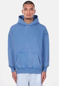 Lyseblå hoodie lavet af blødt stof. Har en frontlomme, ribbede manchetter og diskret logo-broderi på brystet.