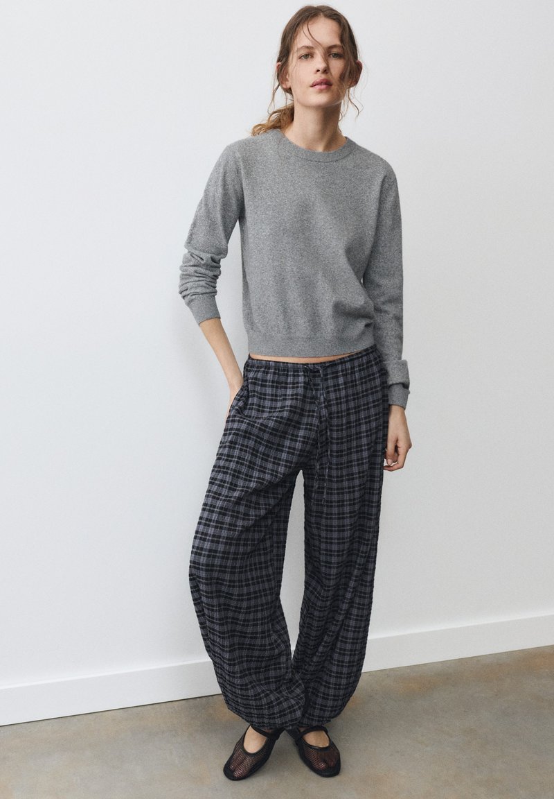 Jeune femme debout, une main dans la poche, portant un pull gris à manches longues, un pantalon large à carreaux foncés et des chaussures noires en résille devant un mur uni.