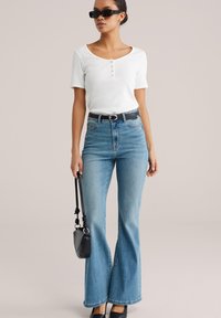 Top alb cu mâneci scurte și detalii cu nasturi, combinat cu blugi evazați bleu deschis, cu talie înaltă. Accesoriile includ o curea neagră și o geantă de mână.