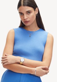 Top blu ribbed senza maniche, collane d'oro a strati con pendenti a forma di cuore, bracciale in argento e un orologio con quadrante bianco e cinturino beige.
