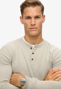 Lichtgrijze lange mouw henley-shirt met drie bruine knopen bij de kraag. Een tan leren horloge met een blauwe wijzerplaat om de pols.