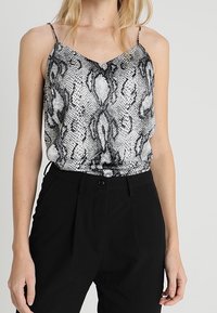 Svart och vit ormskinnsmönstrad linne med tunna band och V-ringning, bärs med höga svarta byxor. Slät textur, lättviktsmaterial.