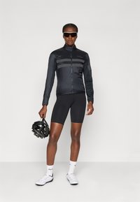 Sort cykeljakke med grå striber, letvægtsstof, lynlås foran. Bæres med sorte shorts, hvide cykelsko, holder en sort hjelm.