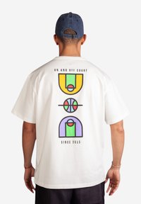 Bílý bavlněný tričko s barevnými geometrickými grafickými prvky souvisejícími s basketbalem na zadní straně, s texty "NA HŘIŠTI I MIMO NĚJ" a "OD ROKU 2013".