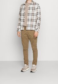 Koszula w kratę w kolorach białym, beżowym i granatowym z zapięciem na guziki; beżowy t-shirt z okrągłym dekoltem; brązowe spodnie cargo; jasnoszare trampki.