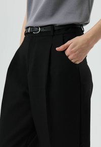 Pantalon noir avec plis, en tissu lisse. Doté d'une ceinture noire avec une boucle argentée, et de poches latérales visibles.