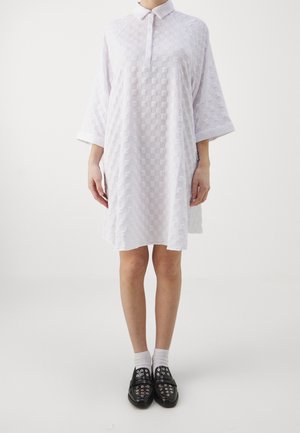 Nainen, jolla on valkoinen, teksturoidusta kankaasta valmistettu, polvipituinen mekko, jossa on kaulus ja leveät hihat, valkoiset sukat ja mustat kengät metallisilla silmukoilla.