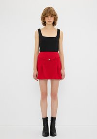 Top sin mangas negro de escote cuadrado combinado con una falda roja ajustada y plisada que presenta un cinturón con detalle dorado. Lleva botines negros.}}