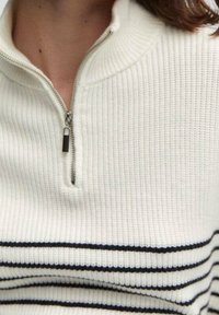 Sweter zapinany na suwak w kremowym kolorze z prążkowaną fakturą, zdobiony granatowymi poziomymi paskami i krótkim kołnierzem. Metalowy zamek przy szyi.