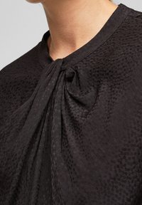 Blusa nera con tessuto testurizzato e un motivo sottile, scollatura drappeggiata e morbide pieghe al colletto.
