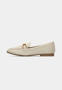 Mocassin beige pour femme avec bout pointu, boucle en métal doré, petit talon en bois et surpiqûres le long du bord supérieur.