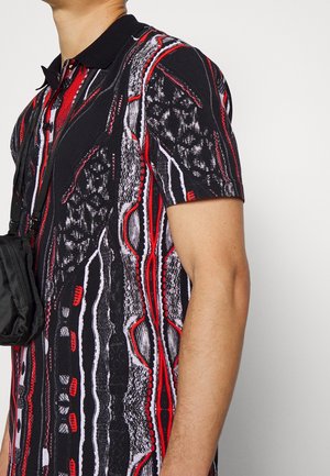 Mężczyzna w czarnej, czerwonej i białej koszulce polo z krótkim rękawem o wzorze, noszący małą czarną torbę na pasku przez ramię.