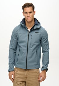 Veste softshell bleu clair avec fermeture éclair à l'avant, poches latérales et capuche, avec un intérieur en denim bleu foncé et un détail de logo discret.