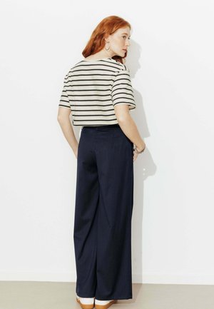 Femme aux cheveux roux portant un haut court rayé noir et blanc, un pantalon large bleu marine, des chaussures blanches, debout de profil contre un mur blanc.