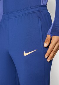 Modré športové nohavice s elastickým pásom a bočnou vreckom, s oranžovým logom Nike. Hladká, ľahká tkanina.
