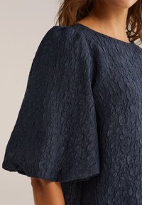 Tumši zila teksturēta blūze ar strukturētām, pūkainām piedurknēm un apaļu kakla izgriezumu, izgatavota no nedaudz krunkainas auduma.