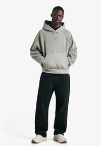 Világoszöld kapucnis pulóver elülső zsebbel, bordázott mandzsettákkal és logóval. Fekete, laza szabású nadrággal és fehér tornacipővel párosítva.