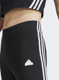 Černé sportovní legíny s třemi bílými pruhy běžícími po bocích a malým bílým logem Adidas blízko pasu.