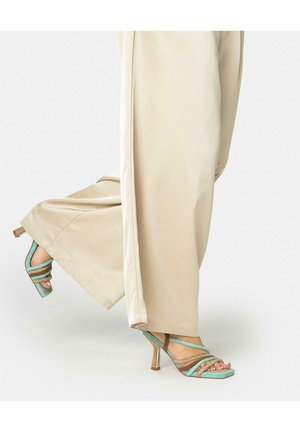 Gambe con pantaloni beige a gamba larga e sandali con cinturini turchese e marroni con tacco alto, un piede leggermente sollevato.