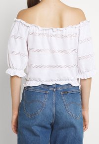 Bílý top bez ramínek s krajkovými detaily a volánkovými okraji, kombinovaný s vysokými modrými džíny s zakřivenými zadními kapsami.