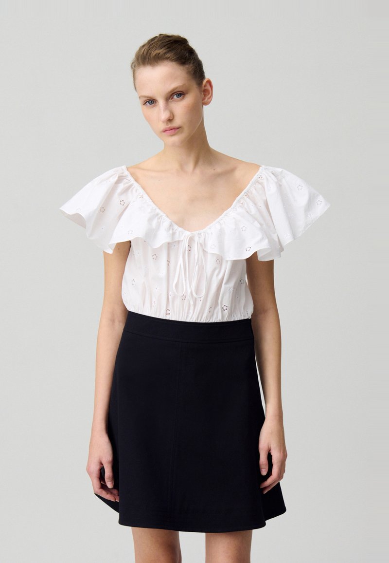 Blouse blanche avec des manches volantes et des détails floraux découpés, associée à une mini-jupe noire évasée, dotée d'une taille cintrée et d'une texture lisse.
