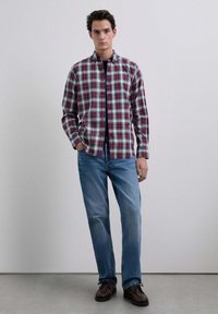 Giovane che si trova contro un muro semplice, indossando una camicia a quadri rossa e grigia, jeans blu e scarpe stringate marroni scure.