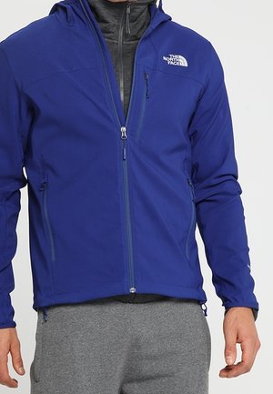 Veste zippée bleue North Face portée par-dessus un sweat à capuche zippé gris foncé, associée à un pantalon de survêtement gris sur une personne debout.