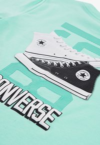 Teal T-skjorte med grafikk av svarte og hvite Converse høy-top sneakers, med "CONVERSE" trykket fremtredende i fet svart typografi.