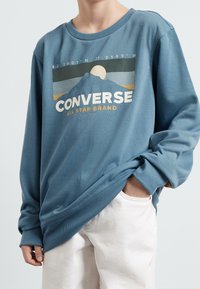 Person wearing blue long-sleeve Converse sweatshirt with mountain and sun graphic, white pants, hand in pocket, against plain background.