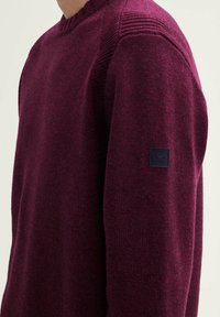 Pull en maille bourgogne avec un motif texturé sur les épaules, encolure ronde et un écusson noir sur la manche.