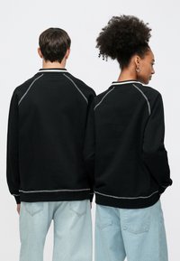 Μαύρο φούτερ με αντίθετη λευκή ραφή, ριμπ γιακά και μανίκια ράγκλαν, σε συνδυασμό με ανοιχτόχρωμο τζιν παντελόνι.