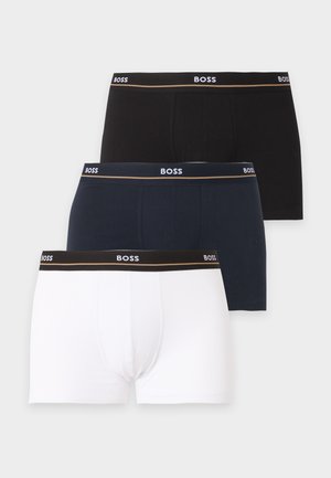 Uomo atletico dalla pelle scura in piedi a piedi nudi che indossa boxer bianchi con fascia nera in vita su sfondo chiaro e uniforme.